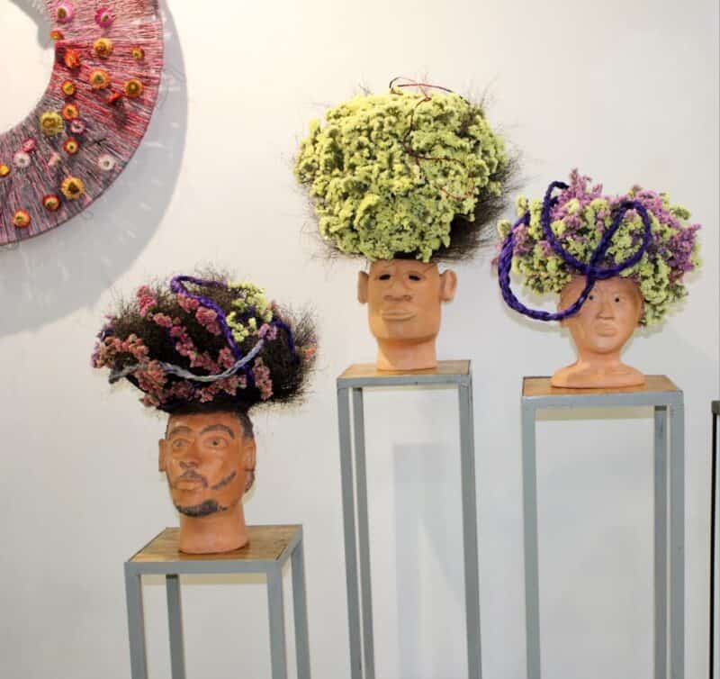 Three clay heads stand on slim metal plinths, each with a different face and a towering headpiece of dried flowers. Sprays of pink, lilac and pale-yellow blooms mix with dark grasses and looping purple braids at the Roedean Art Fair in Houghton on October 1, 2026. Photo: Naziya Davids-Easthorpe
