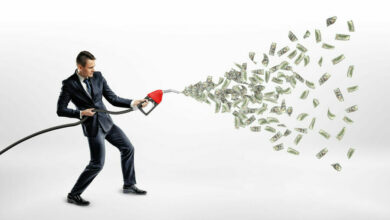 A smiling businessman holding a gas nozzle while many dollar bills are flying out of it instead of petrol.