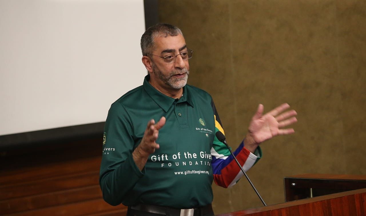 man speaking in front of a microphone, gesturing with both his hands