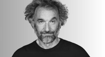 A black and white headshot of a middle-aged man with a beard and curly hair, looking directly at the viewer with a slight smile