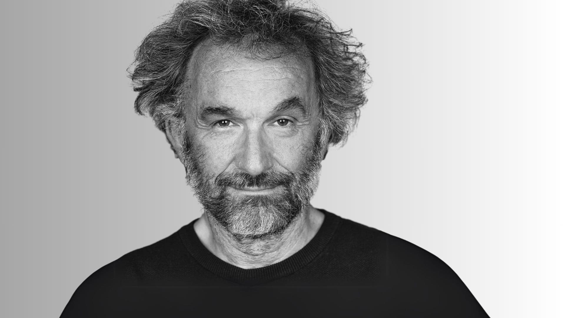 A black and white headshot of a middle-aged man with a beard and curly hair, looking directly at the viewer with a slight smile