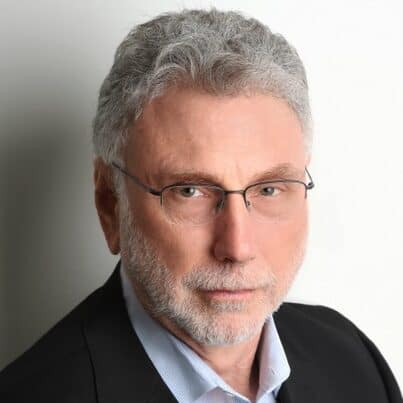 A headshot of a middle-aged man with gray, curly hair and a gray beard, wearing glasses and a dark suit jacket over a light blue collared shirt. He is looking directly at the viewer.