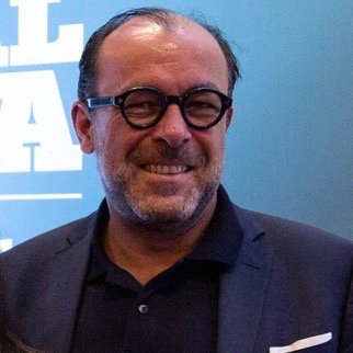 A smiling middle-aged man with a receding hairline, wearing round black glasses, a navy blazer, and a dark shirt, standing in front of a blue background with white text.