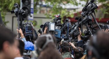 professional video cameras, many mounted on tripods, to capture an event. The cameras and the backs of people's heads dominate the foreground, suggesting a focus on media coverage or a public gathering of significant interest.