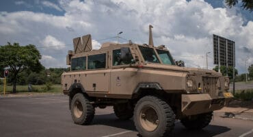 A beige SANDF military armored vehicle with large tires is parked on a road under a blue sky with fluffy clouds. Trees and a traffic light are in the background.