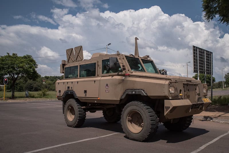 A beige SANDF military armored vehicle with large tires is parked on a road under a blue sky with fluffy clouds. Trees and a traffic light are in the background.
