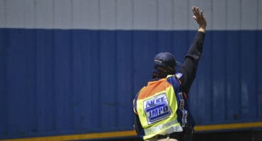 A JMPD officer pictured, extending their arm into the air, gesturing for a car to stop. The officer is seen wearing a JMPD vest and a blue cap.