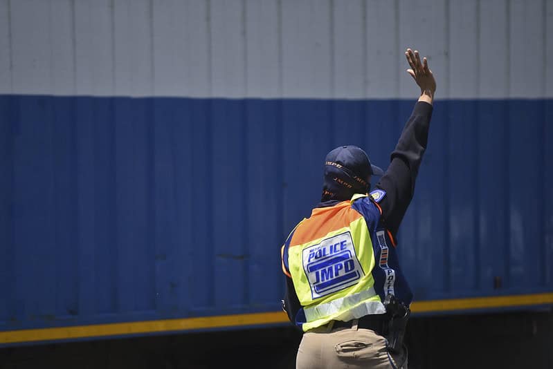 A JMPD officer pictured, extending their arm into the air, gesturing for a car to stop. The officer is seen wearing a JMPD vest and a blue cap.