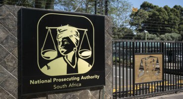 The entrance of the National Prosecuting Authority (NPA) in Pretoria is seen 22 April 2024. Sign for the National Prosecuting Authority of South Africa on a stone wall. It features Lady Justice and scales. A gate and trees are in the background.