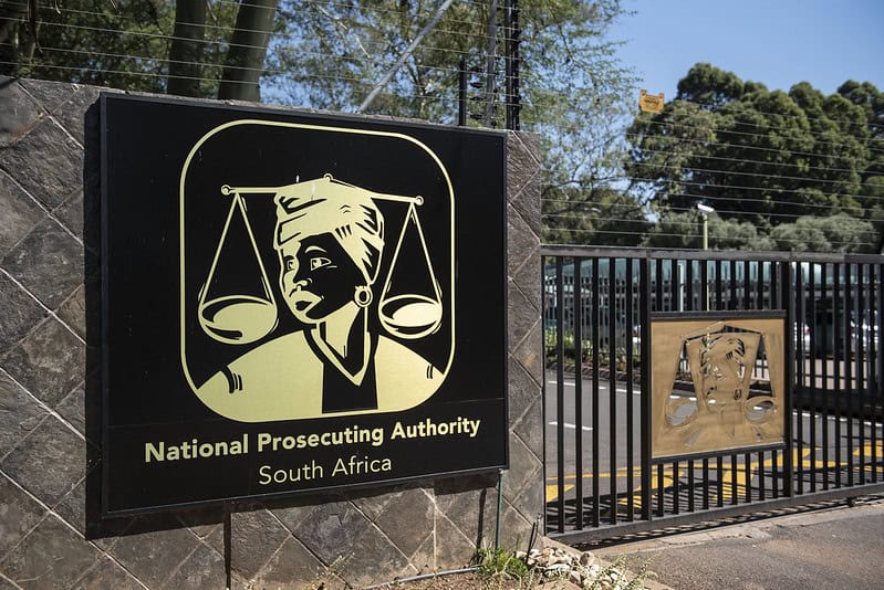 The entrance of the National Prosecuting Authority (NPA) in Pretoria is seen 22 April 2024. Sign for the National Prosecuting Authority of South Africa on a stone wall. It features Lady Justice and scales. A gate and trees are in the background.