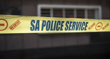 Yellow "SA Police Service" tape with "No Entry" symbols stretches across a blurred urban background, conveying caution and restricted access.