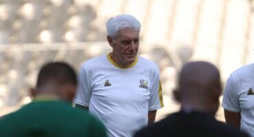 Hugo Broos in a white sports shirt stands outdoors with three people in the foreground, out of focus.