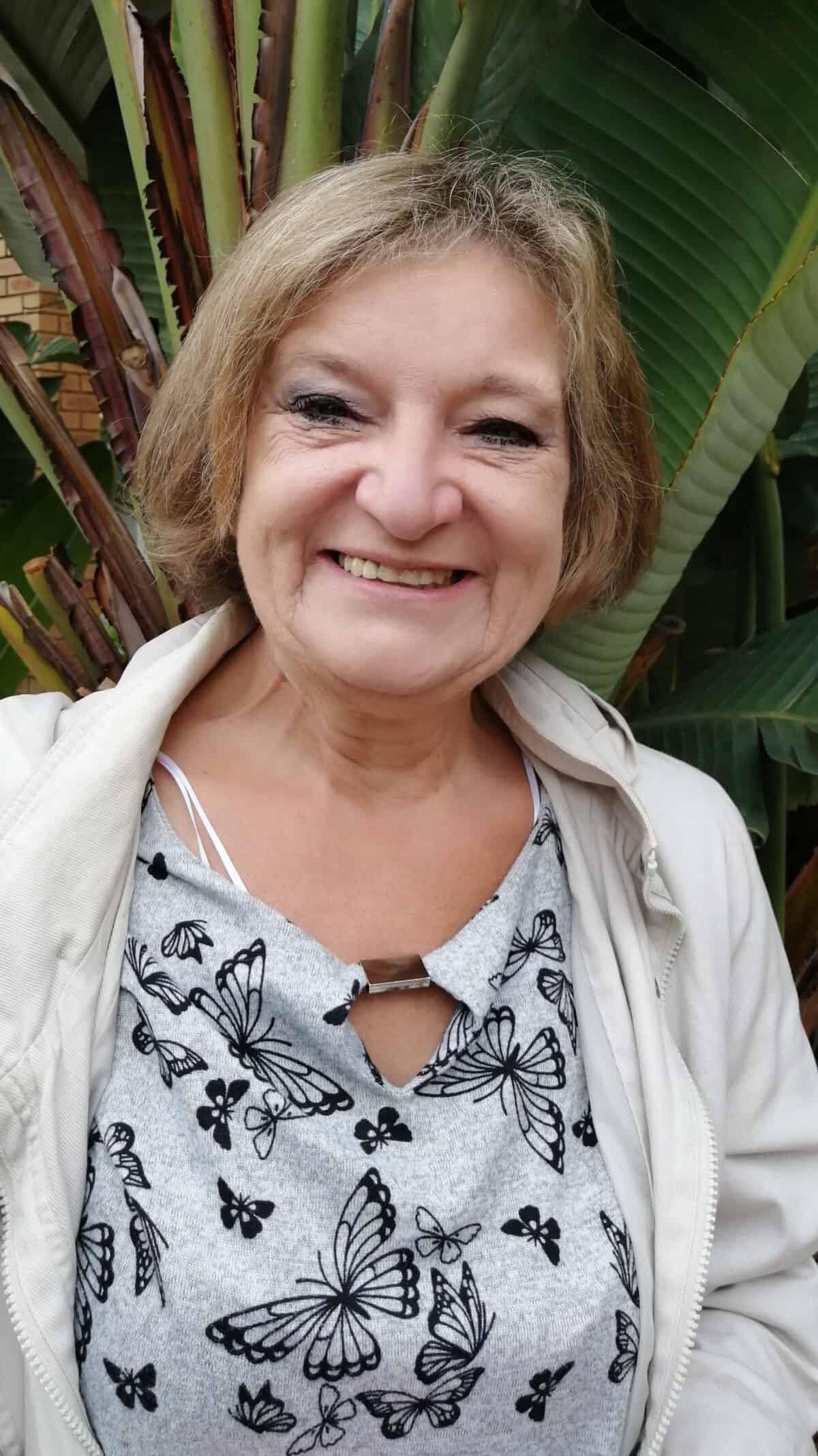 A smiling caregiver or therapist stands outdoors in front of green tropical leaves, wearing a light jacket and a butterfly-patterned top, symbolising positivity and resilience in mental health care.