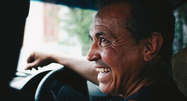 Smiling man with short dark hair, seated in a car, looking to the side. His expression is joyful, creating a warm and relaxed atmosphere.
