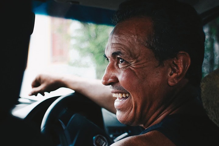 Smiling man with short dark hair, seated in a car, looking to the side. His expression is joyful, creating a warm and relaxed atmosphere.