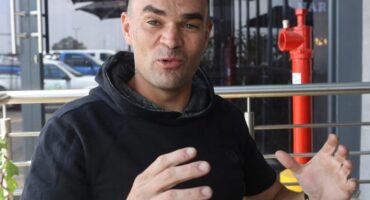 Man gesturing while speaking during an interview, wearing a black hoodie, with an urban setting in the background.