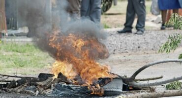 A tire and debris are burning on the ground, producing thick black smoke – a stark sign of dysfunction in a failed state – as several people stand in the background.