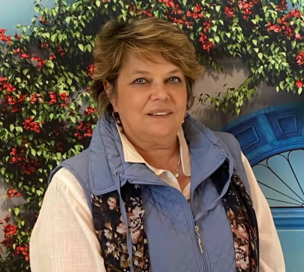 A caregiver or healthcare professional stands smiling in front of a floral backdrop, wearing a light blue quilted vest and white shirt, representing dedication and compassion in elder care.
