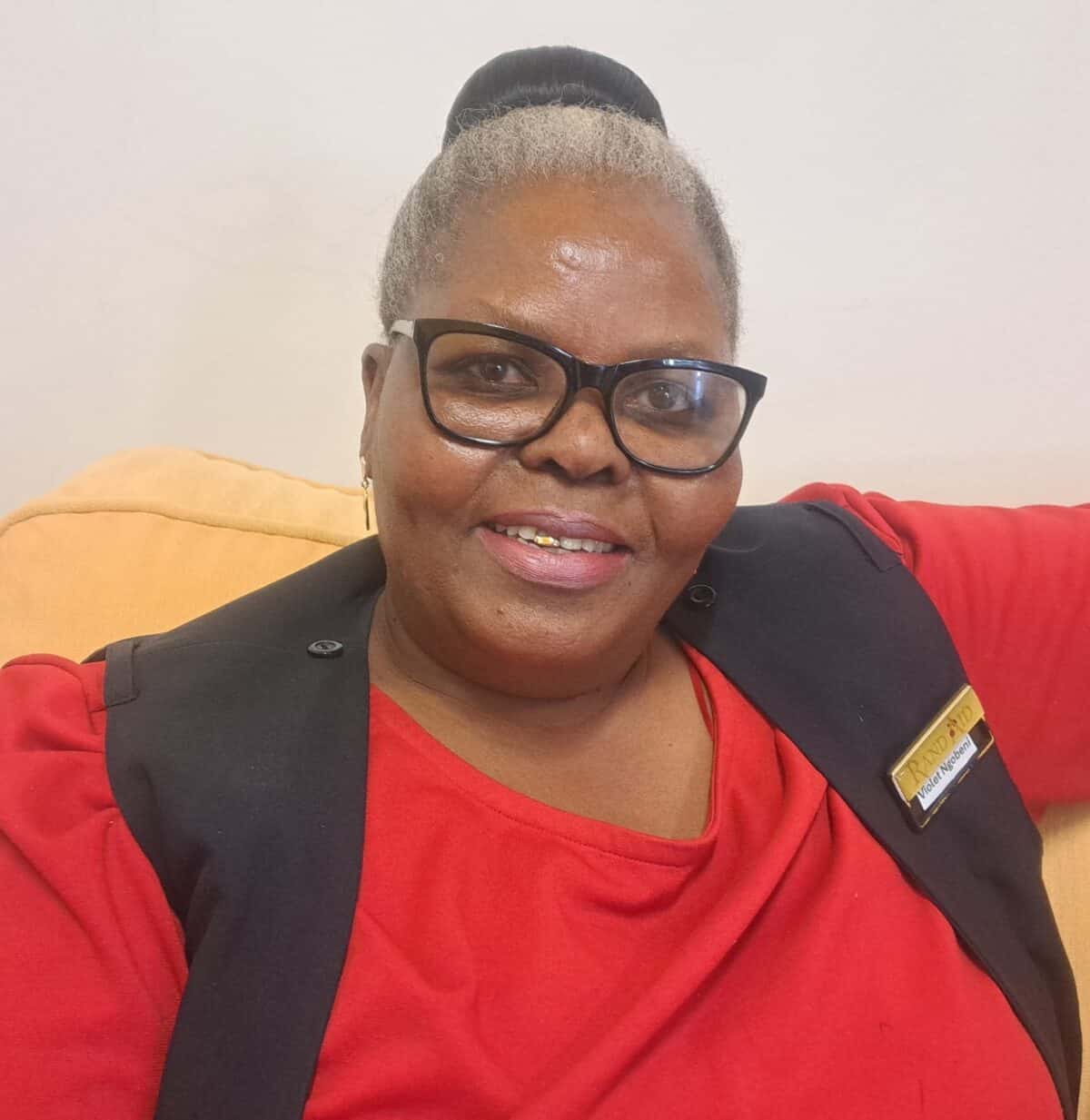 Black woman with grey hair pulled back in a wearing dark rimmed glasses sitting on a beige couch smiling at the camera.