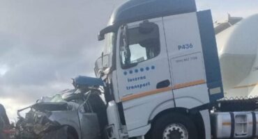 A damaged silver car is crushed beneath the front of a large white truck labeled "lucerne transport," under a cloudy sky, conveying a sense of destruction.