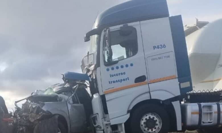 A damaged silver car is crushed beneath the front of a large white truck labeled "lucerne transport," under a cloudy sky, conveying a sense of destruction.