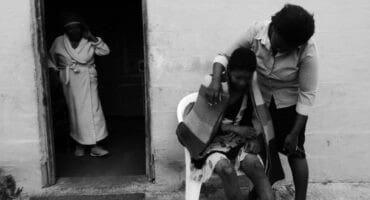 A black and white image depicts a somber scene. A woman leans over, comforting another wrapped in a blanket, seated outside a doorway. Another figure stands in the background, watching.