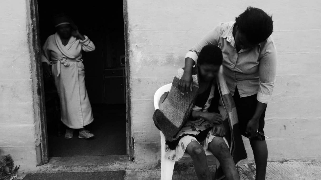 A black and white image depicts a somber scene. A woman leans over, comforting another wrapped in a blanket, seated outside a doorway. Another figure stands in the background, watching.