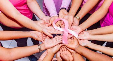 A diverse group of people in pink shirts form a circle with hands touching, holding a pink ribbon in the centre, symbolising unity and breast cancer awareness.