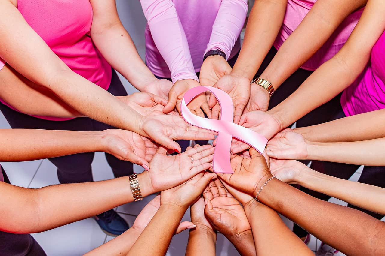 A diverse group of people in pink shirts form a circle with hands touching, holding a pink ribbon in the centre, symbolising unity and breast cancer awareness.