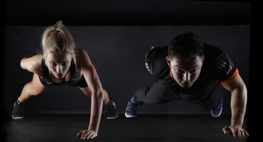 A man and woman perform push-ups side by side on a dark floor. Both are focused and determined, conveying strength and fitness. The scene is lit dramatically.