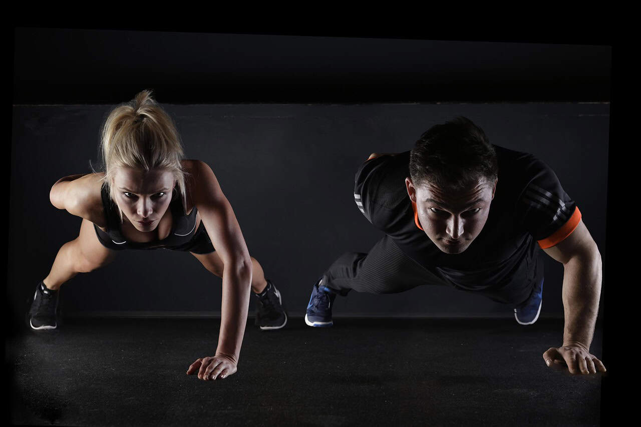 A man and woman perform push-ups side by side on a dark floor. Both are focused and determined, conveying strength and fitness. The scene is lit dramatically.