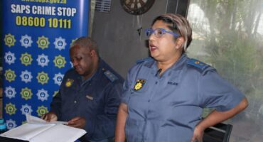 Two police officers in uniform are seen in an indoor setting. One is reading documents at a table, while the other stands speaking. A "SAPS Crime Stop" sign is visible.