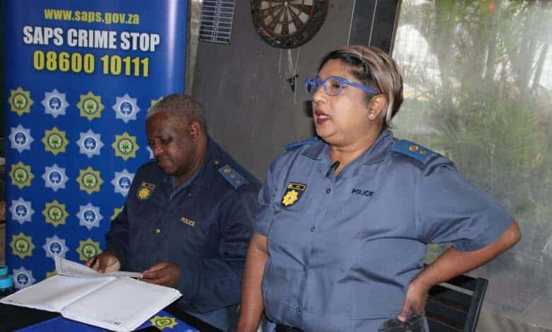 Two police officers in uniform are seen in an indoor setting. One is reading documents at a table, while the other stands speaking. A "SAPS Crime Stop" sign is visible.