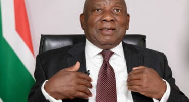 President Cyril Ramaphosa dressed in a suit and tie gestures while speaking, seated in front of a South African flag. The expression is focused, suggesting an official setting.