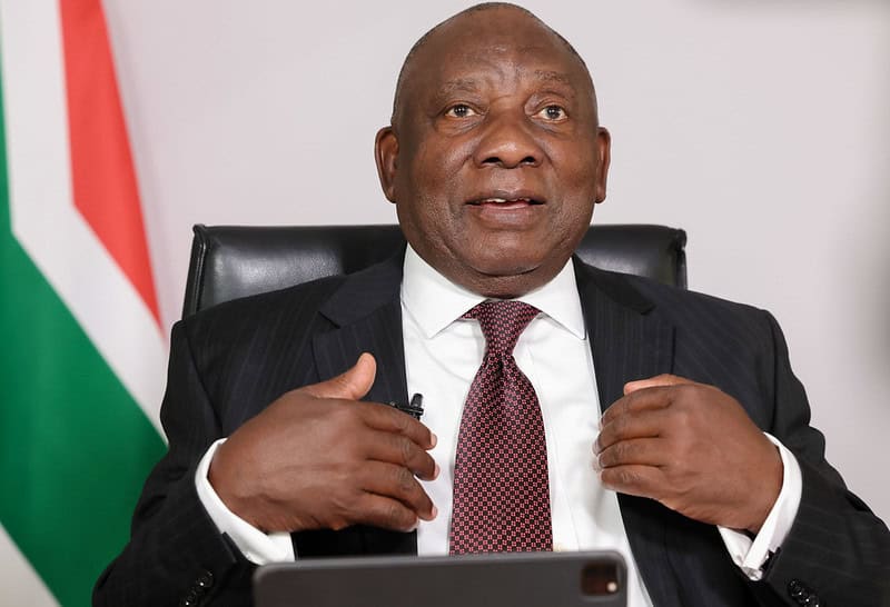 President Cyril Ramaphosa dressed in a suit and tie gestures while speaking, seated in front of a South African flag. The expression is focused, suggesting an official setting.