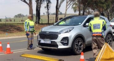 Traffic officers surround a silver car at a traffic stop with traffic cones visible in the foreground.