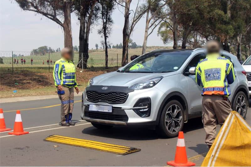 Traffic officers surround a silver car at a traffic stop with traffic cones visible in the foreground.
