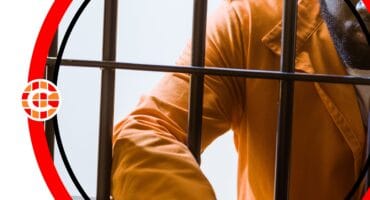 A man in an orange jumpsuit, leaning against bars in a jail cell.