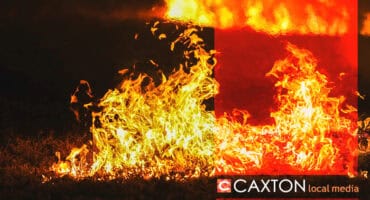 Vibrant orange and yellow flames engulf grass in a dark setting, creating an intense, fiery scene. The bottom right reads "CAXTON local media."