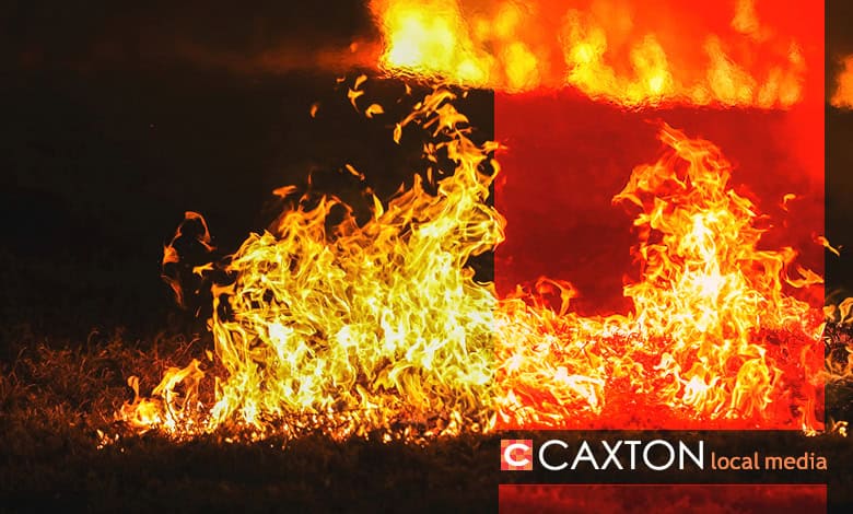 Vibrant orange and yellow flames engulf grass in a dark setting, creating an intense, fiery scene. The bottom right reads "CAXTON local media."