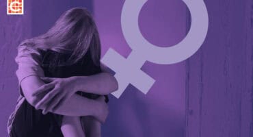 A solitary person sits curled up in a corner with head hidden by hair. A large female gender symbol overlays the image, conveying a sense of vulnerability.