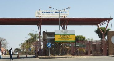 Entrance of Seshego Hospital featuring a large red metal canopy, signage, and lush greenery. The clear sky adds to the calm atmosphere.