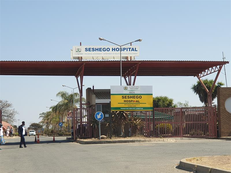Entrance of Seshego Hospital featuring a large red metal canopy, signage, and lush greenery. The clear sky adds to the calm atmosphere.