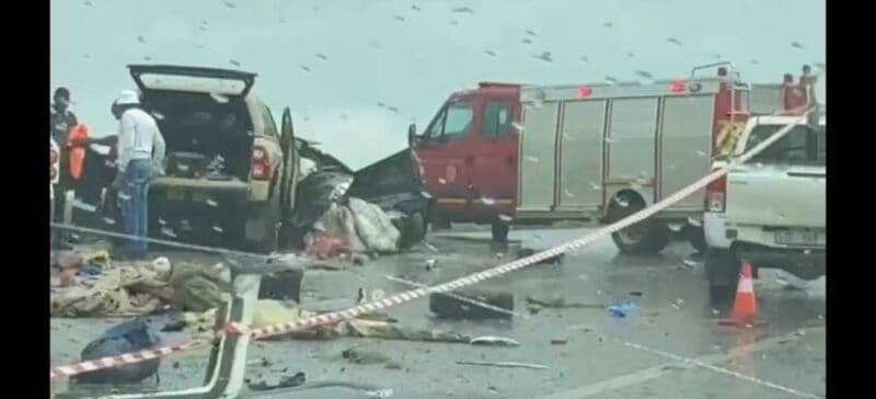 An accident scene depicting a rainy road, with car wrecks, an emergency vehicle and debris covering the road visible.