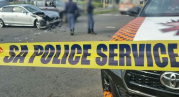 A damaged silver car is visible behind a yellow "SA Police Service" tape. Officers are blurred in the background, suggesting a recent accident scene.