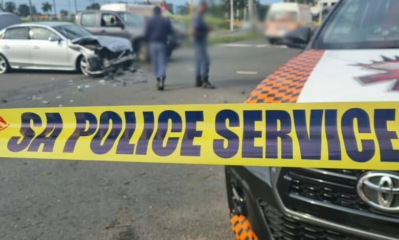 A damaged silver car is visible behind a yellow "SA Police Service" tape. Officers are blurred in the background, suggesting a recent accident scene.