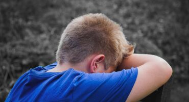 A boy with short blond hair wearing a blue shirt leans his head on his crossed arms in a contemplative pose, suggesting sadness or reflection, against a blurred outdoor background.