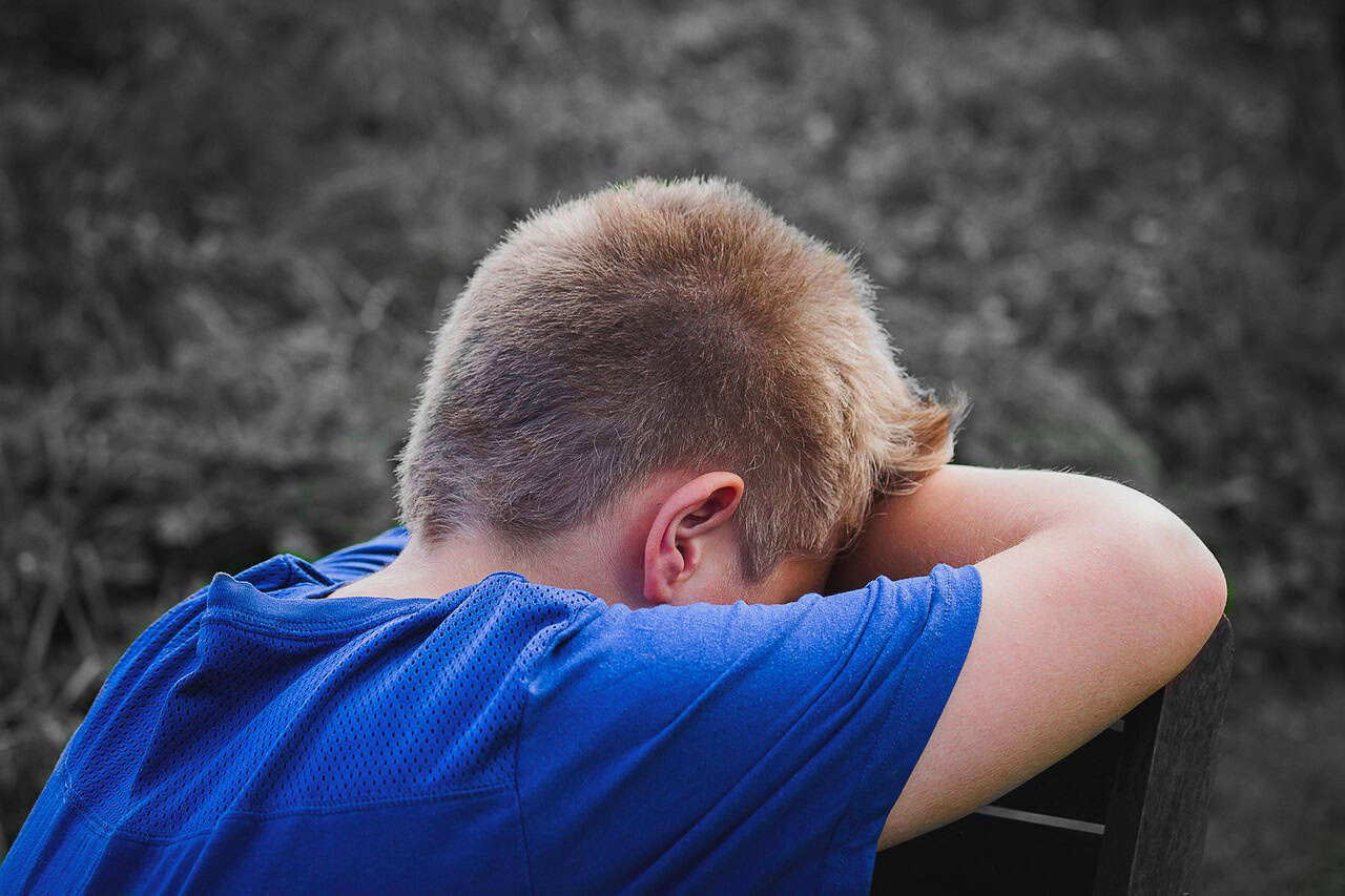A boy with short blond hair wearing a blue shirt leans his head on his crossed arms in a contemplative pose, suggesting sadness or reflection, against a blurred outdoor background.