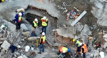 Overhead view of construction workers wearing safety vests and helmets at a site with rubble and a large hole, conveying focus and collaboration.