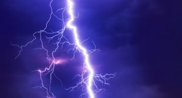 Intense lightning bolt against a dark purple sky, with smaller branching sparks illuminating surrounding clouds, conveying a dramatic and powerful atmosphere.